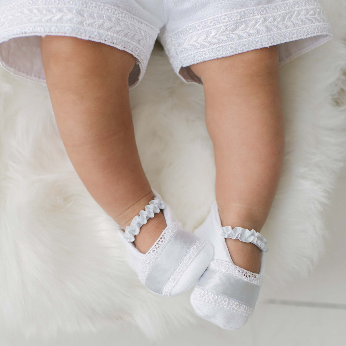 Baby's feet wearing the Henry Booties on a soft white surface. Details show blue ribbon and Venice lace.