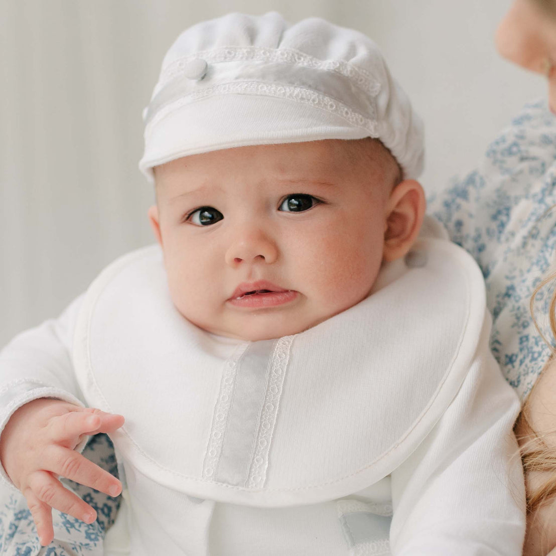 Baby in the Henry Brim Cap and matching 3-Piece Suit and Bib sitting on a woman with blue patterned dress. Part of Baby Beau & Belle's high end baby clothes.