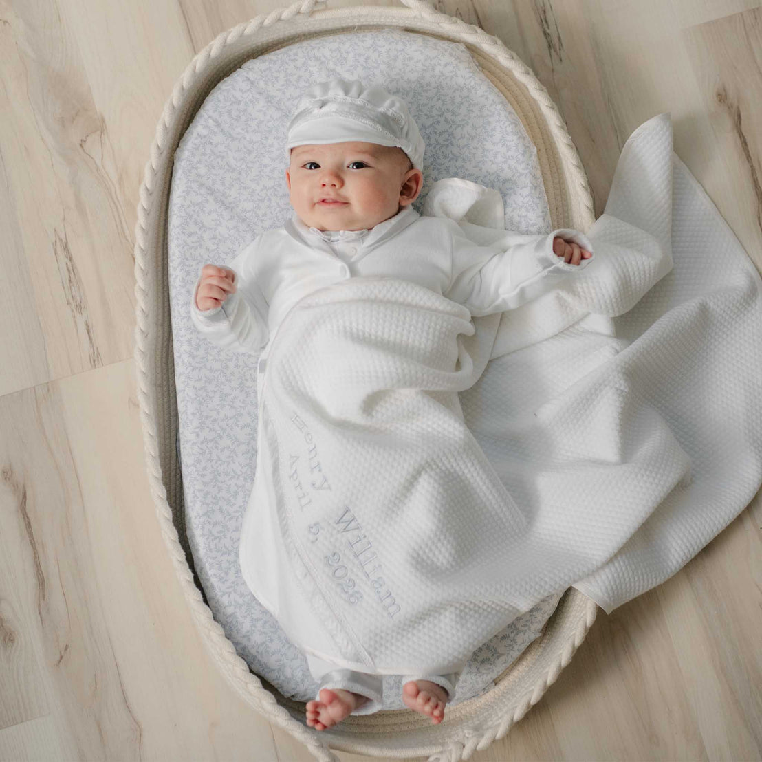 Baby boy wrapped in the Henry Personalized Blanket, lying in a white basket with blue patterned cushion on a wooden floor. Details show the name embroidery on the corner of the blanket. Perfect for unique baby gifts.