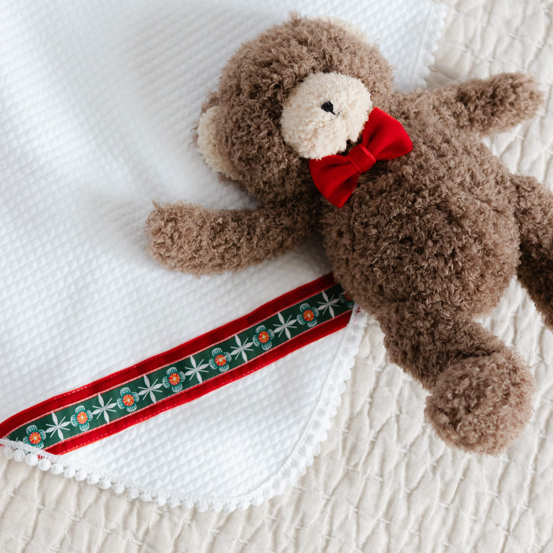 Brown teddy bear with a red bow tie lying on a white quilt blanket