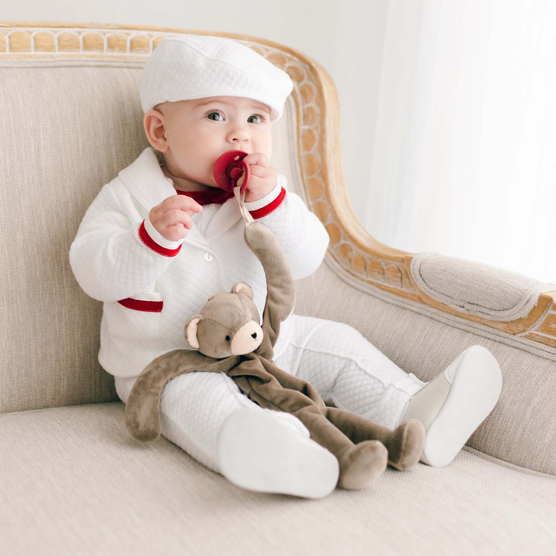 A baby dressed in a white outfit with red accents and a matching hat is sitting on a beige armchair. The little one, holding a pacifier secured with the Silly Bear Buddy | Pacifier Holder, clutches a brown stuffed monkey toy. This ensemble makes for an ideal Christening gift, ensuring the baby stays stylish and safe.