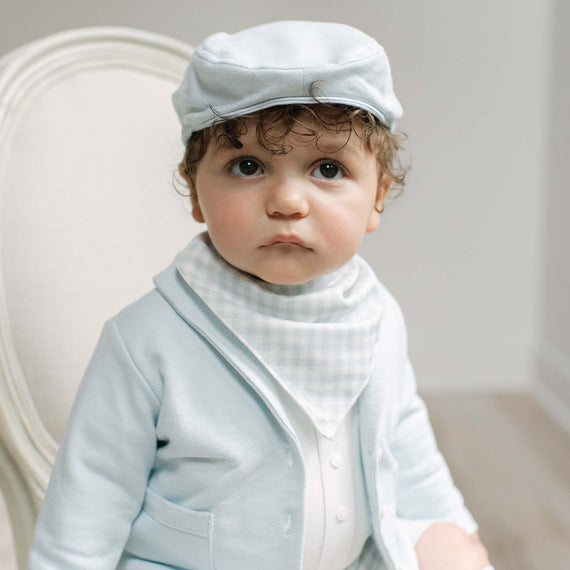 A baby wearing a light blue cap, a light blue cardigan, and a white and light blue gingham bib, sitting in a white upholstered chair.