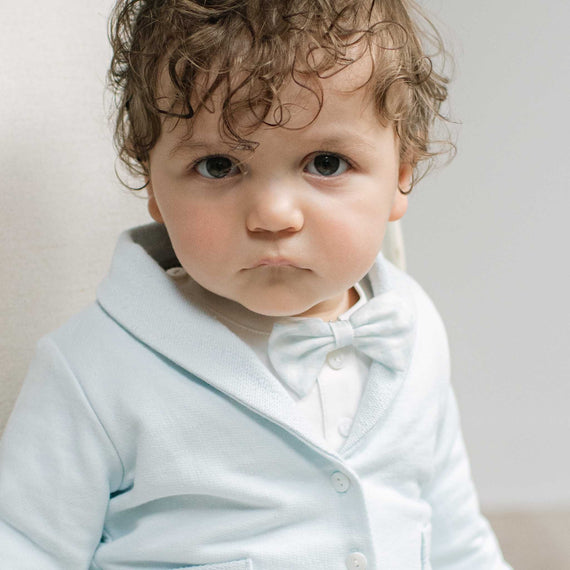 A close-up portrait of a baby with curly brown hair wearing a light blue button-front cardigan with a matching bow tie over a white shirt, set against a soft neutral background.