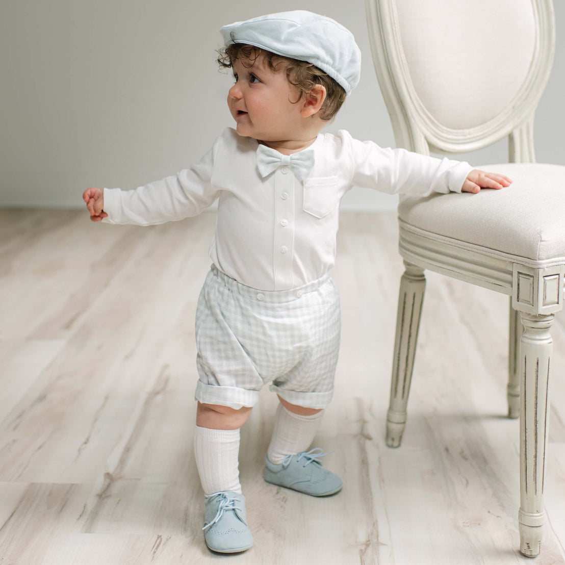 A baby toddler standing on a light wood floor next to a white upholstered chair, wearing a white long-sleeve shirt with a bow tie, white and blue gingham shorts, white socks, and light blue shoes, and a pale blue flat cap.