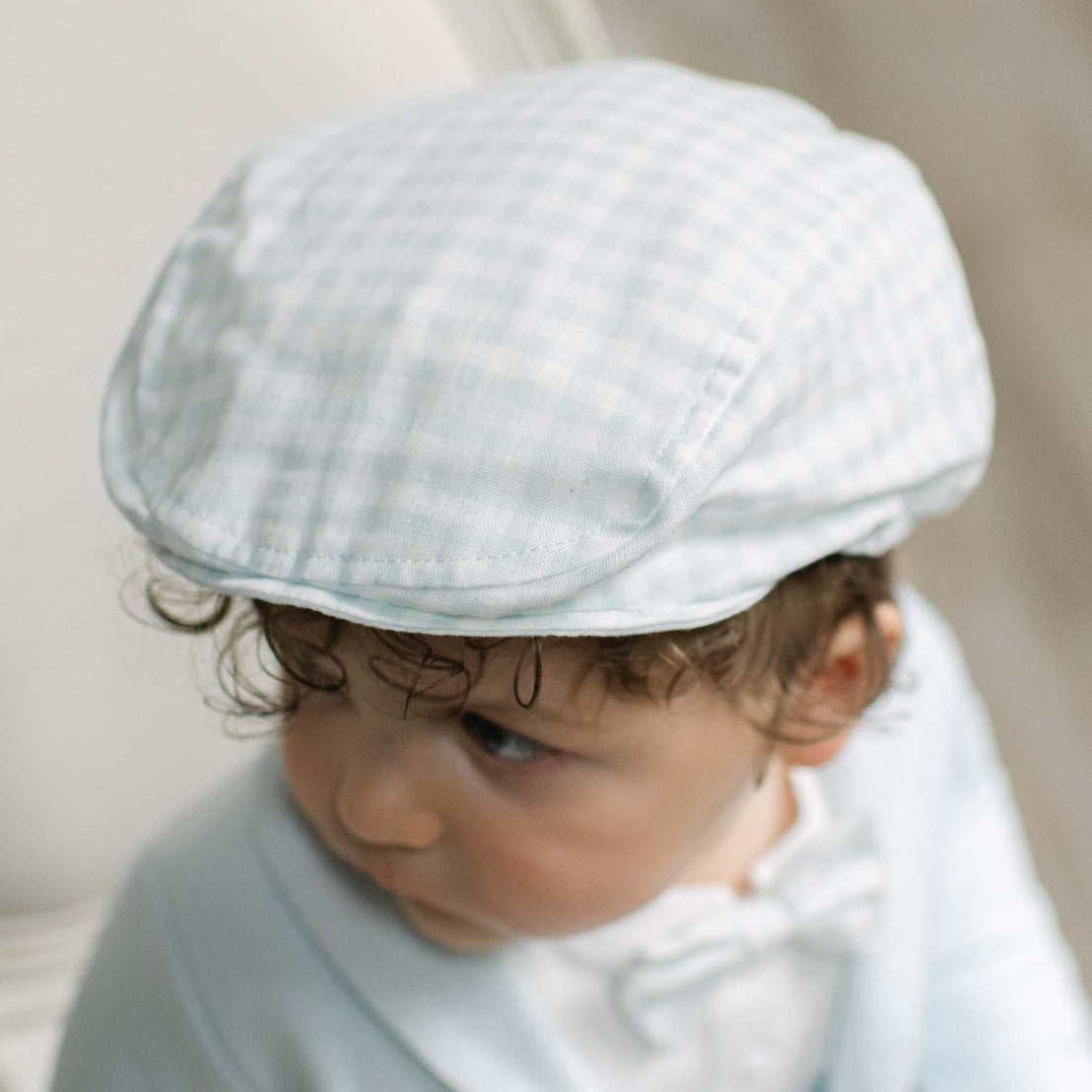 A close-up of a baby wearing a light blue and white gingham check flat cap and a matching light blue top.
