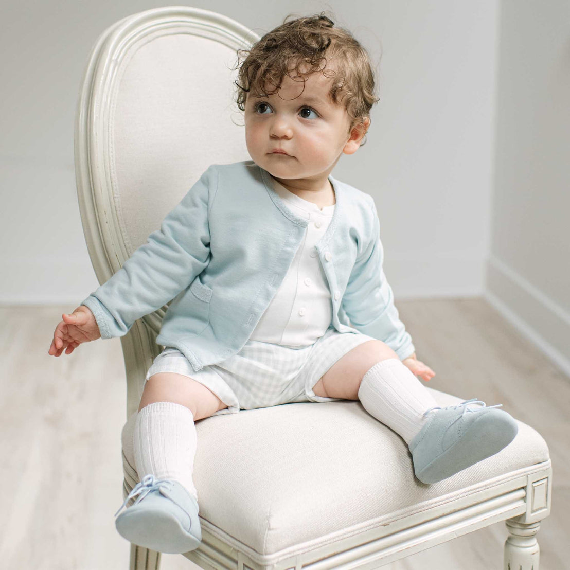 A baby toddler with curly brown hair sits on a white upholstered chair, wearing a light blue cardigan over a white outfit with white socks and pale blue shoes.