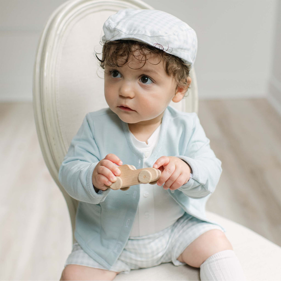A baby sitting on a white upholstered chair wearing a light blue cardigan, matching shorts, a white shirt, white socks, and a white cap, while holding a small wooden toy.