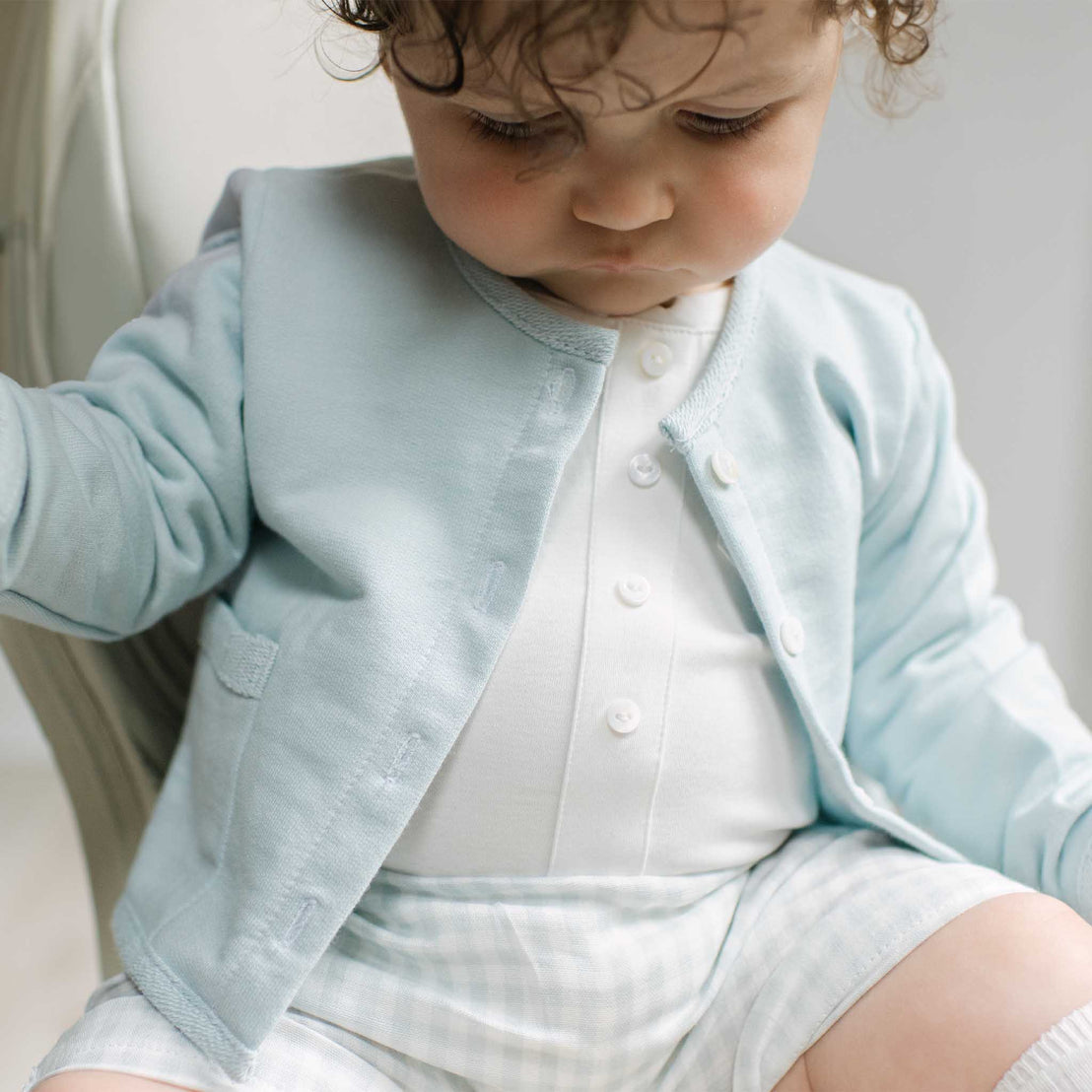 A close-up of a baby wearing a light blue cardigan over a white button-up shirt and white gingham shorts, looking down.