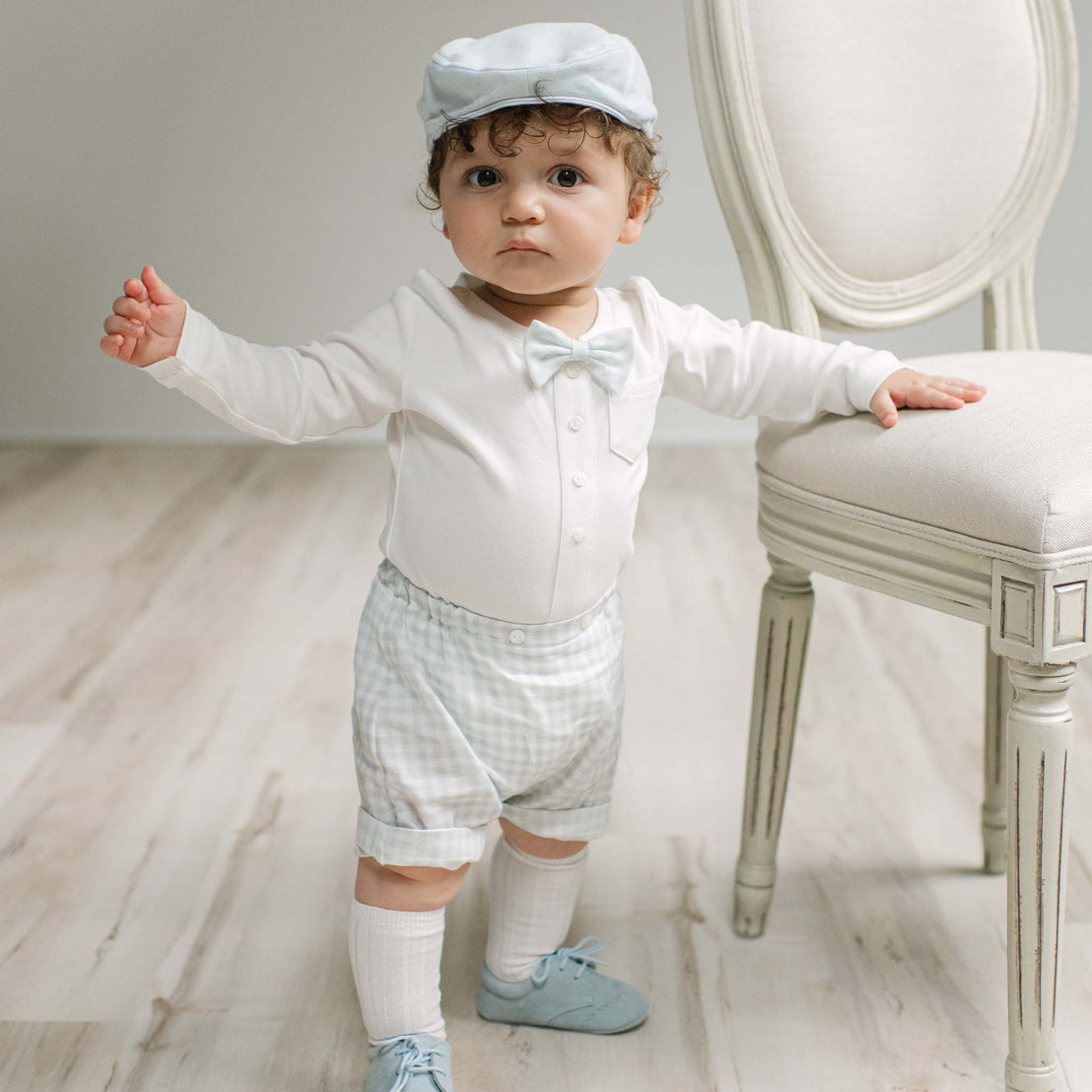 A baby boy wearing a white long-sleeve romper with a bow tie, a white gingham shortalls outfit, a white cap, white socks, and light blue shoes, standing next to a white upholstered chair.