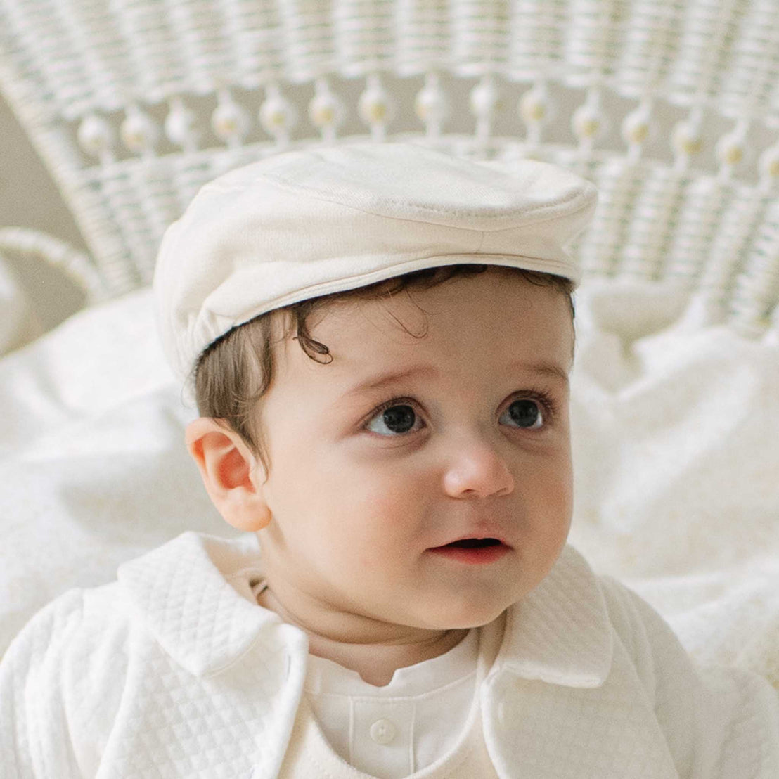 A baby wearing a cream-colored textured cap and matching long-sleeve collared outfit, sitting in a white wicker chair.