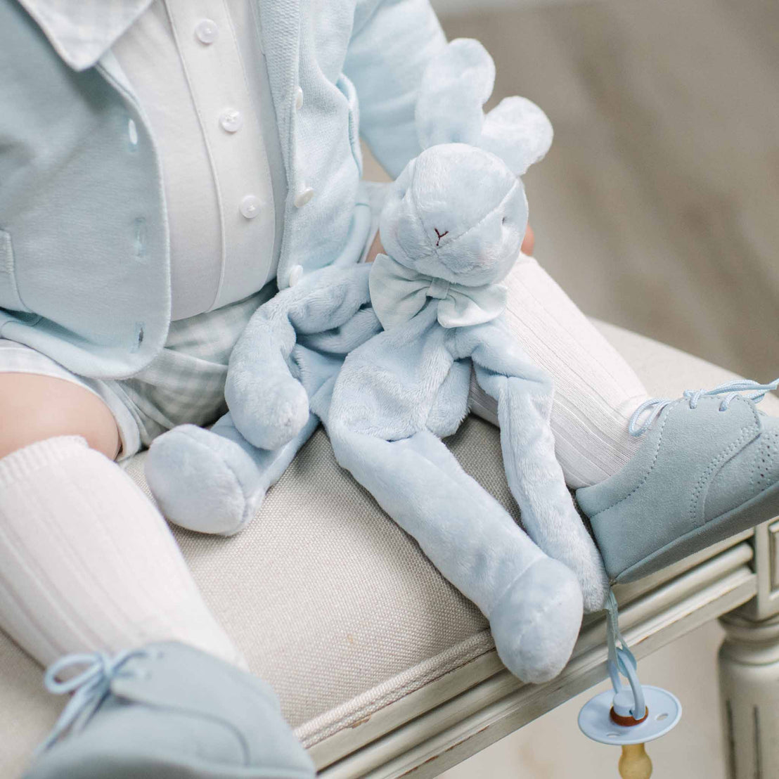 A baby wearing a light blue cardigan, white shirt, white socks, and light blue shoes, holding a matching light blue plush bunny toy while sitting on a beige upholstered bench.