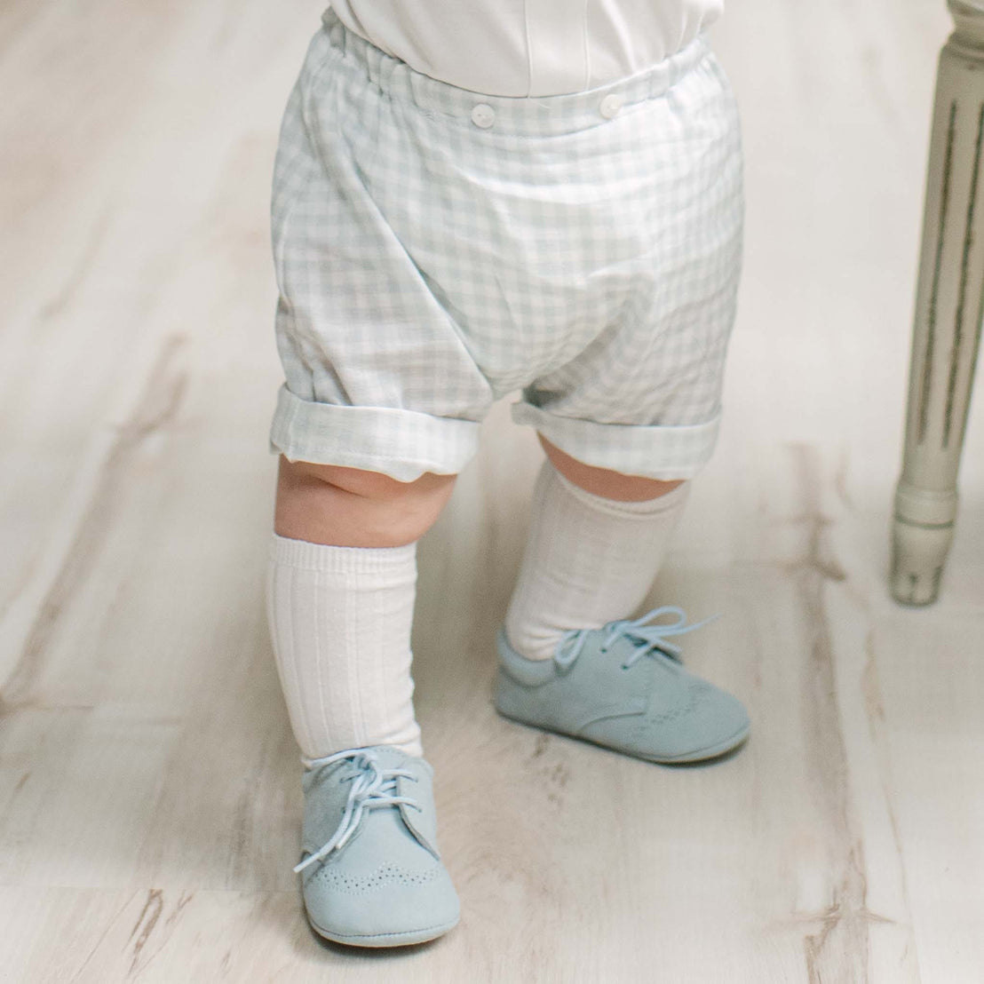 A close-up of a baby's lower body wearing light blue suede shoes, white socks, and pale blue and white gingham check shorts on a light wood floor.