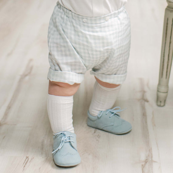 A close-up of a baby's lower body wearing light blue suede shoes, white socks, and pale blue and white gingham check shorts on a light wood floor.