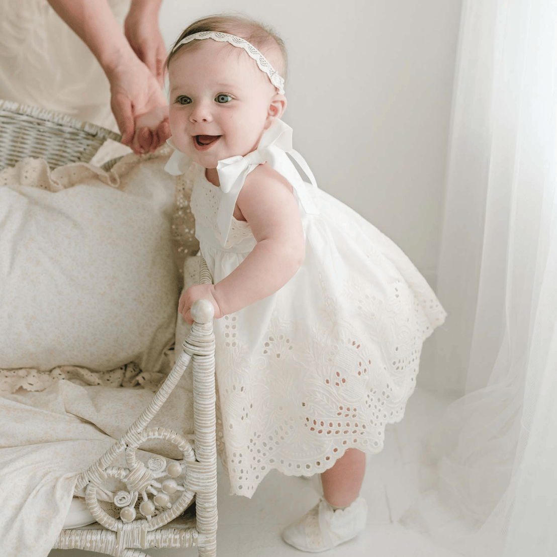 A smiling baby dressed in the Ingrid Romper Dress and matching headband is standing while holding onto a wicker chair, supported by an adult hand behind her. The scene showcases this charming girls' dress, which is handmade in the USA, and is illuminated by warm, natural light.