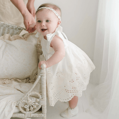 A smiling baby dressed in the Ingrid Romper Dress and matching headband is standing while holding onto a wicker chair, supported by an adult hand behind her. The scene showcases this charming girls' dress, which is handmade in the USA, and is illuminated by warm, natural light.