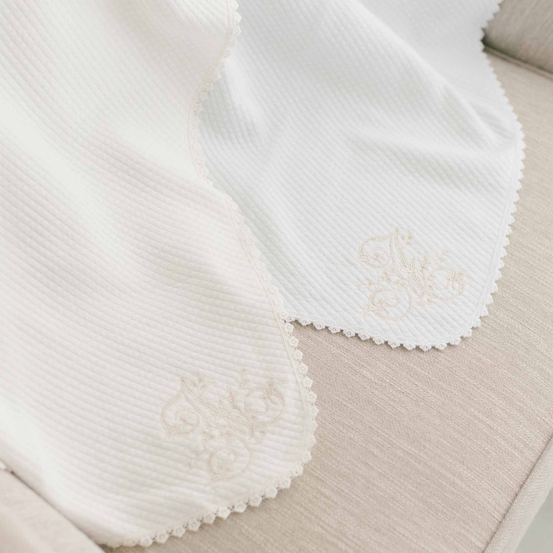 Two white, embroidered  baby blankets with decorative lace edges and textured details, elegantly displayed on a neutral beige surface, featuring letter embroidery personalization.