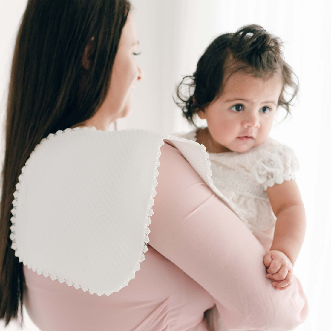 A woman with long brown hair, wearing a pink top, holds a baby dressed in a white outfit adorned with delicate ivory lace. The woman has an Ivory Burp Cloth draped over her shoulder as they stand in a softly lit room.