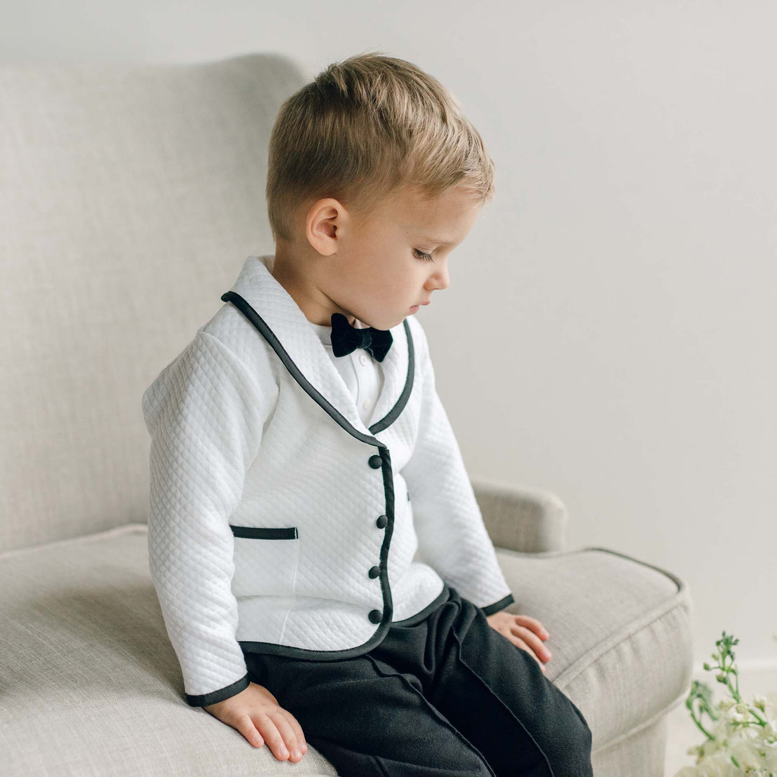 A young boy seated to the side, wearing the James 3-Piece Suit in white and black with matching bow tie.