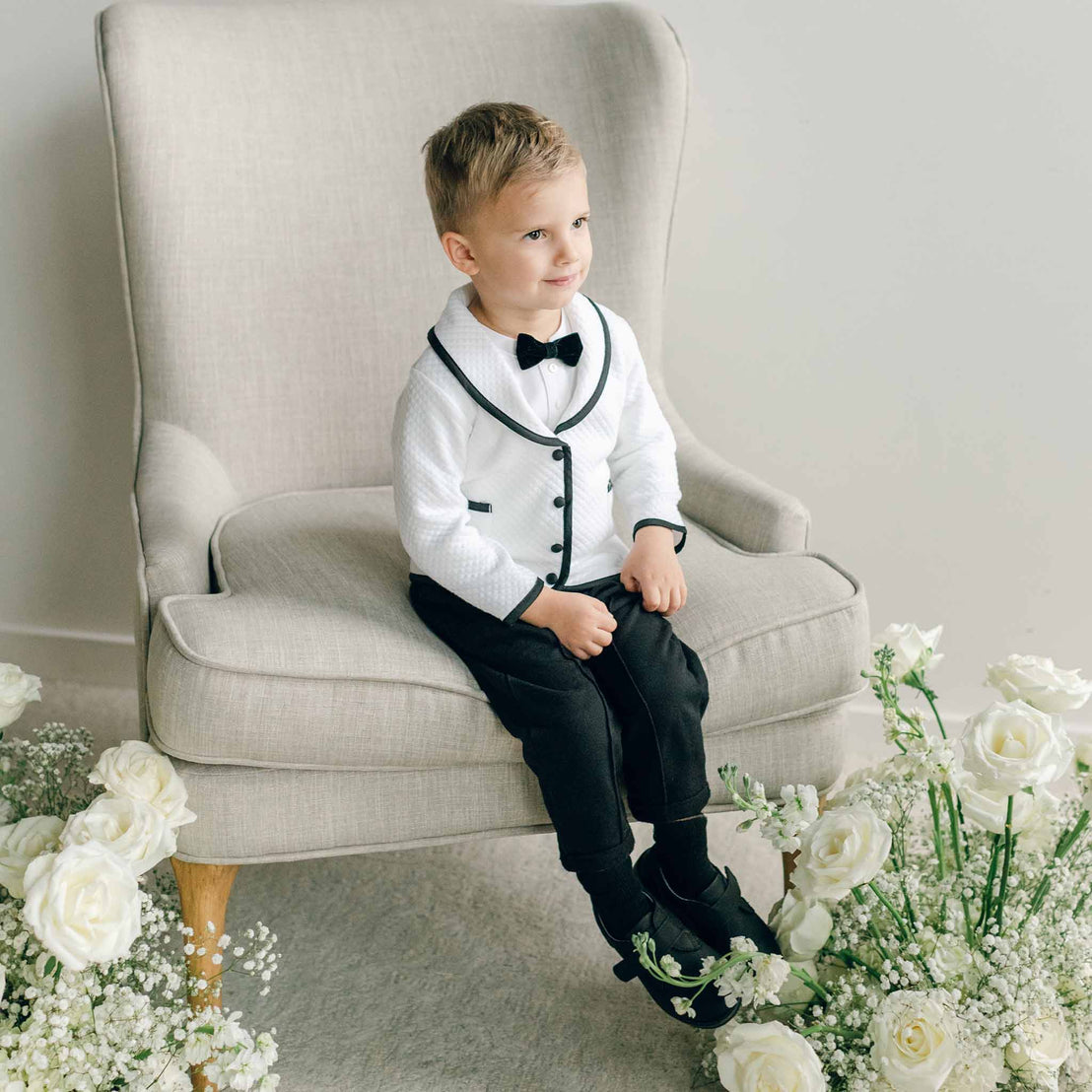 A young boy seated in a light armchair, dressed in the James 3-Piece Suit with a white jacket and coordinating black pants.