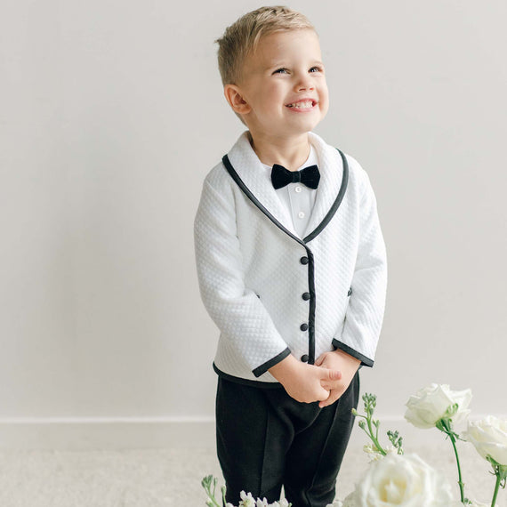 A young boy wearing the James 3-Piece Suit, featuring a white quilted jacket with black trim, black pants, and a black velvet bow tie.