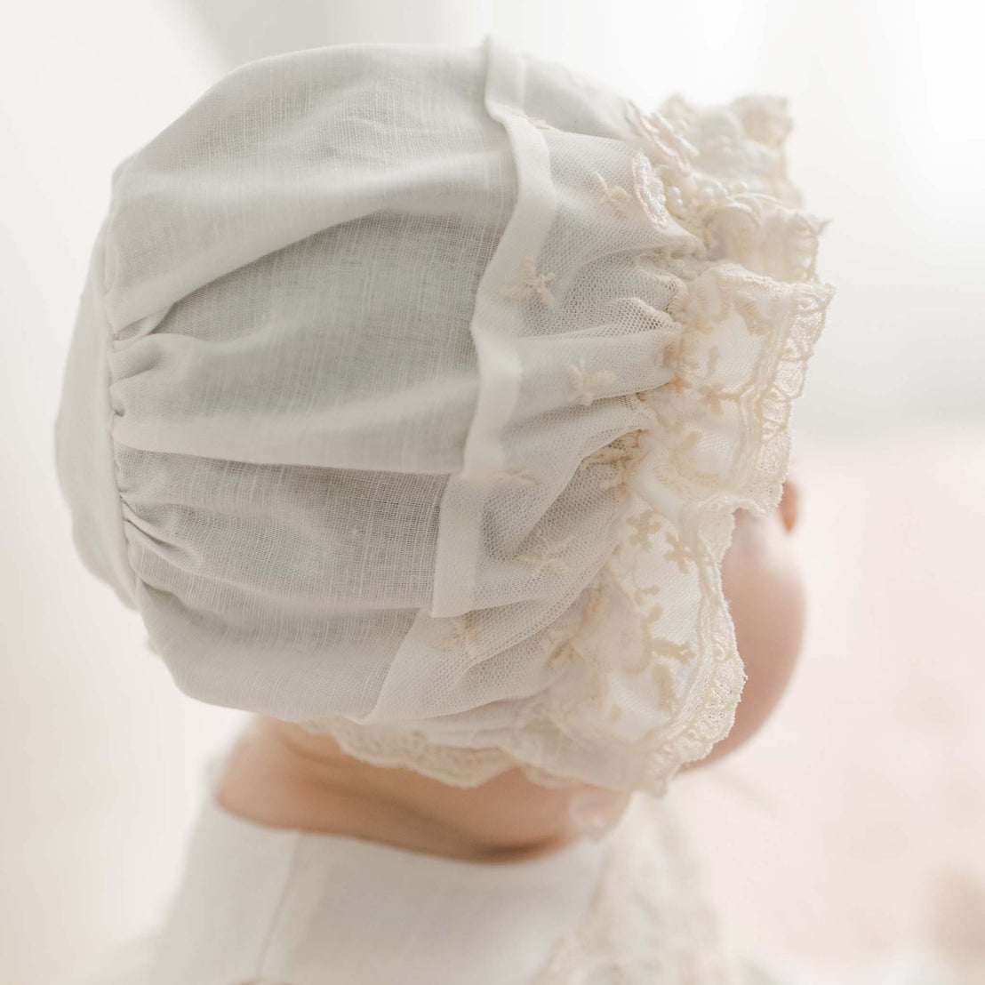The Jessica Linen Bonnet, crafted in a delicate, vintage style with Champagne Lace and soft fabric, elegantly covers the baby's head. A softly blurred background enhances the gentle and serene atmosphere surrounding this beautiful bonnet.