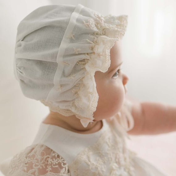 A baby gazes to the side wearing a Jessica Linen Bonnet along with a delicate, vintage-style dress, beautifully detailed with champagne lace and embroidery. This ensemble creates a soft, nostalgic look against a light, airy background.
