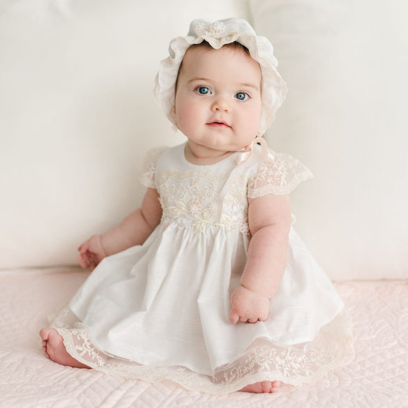 A baby wearing the Jessica Linen Dress & Bloomers, featuring delicate lace details and flutter sleeves, sits on a soft pink surface. The vintage charm of her ensemble enhances her neutral expression against a light, neutral background.