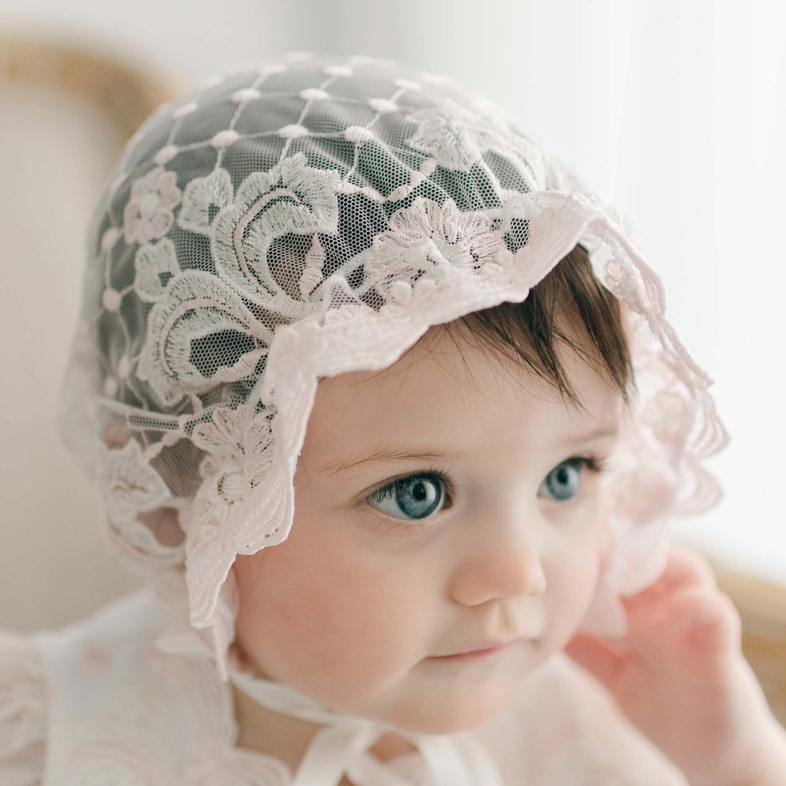 Close-up of a baby girl wearing the Jolie Sheer Lace Bonnet, featuring delicate floral embroidery and soft ruffled edges. The bonnet is tied under her chin with silk ribbons, framing her face as she gazes off to the side.