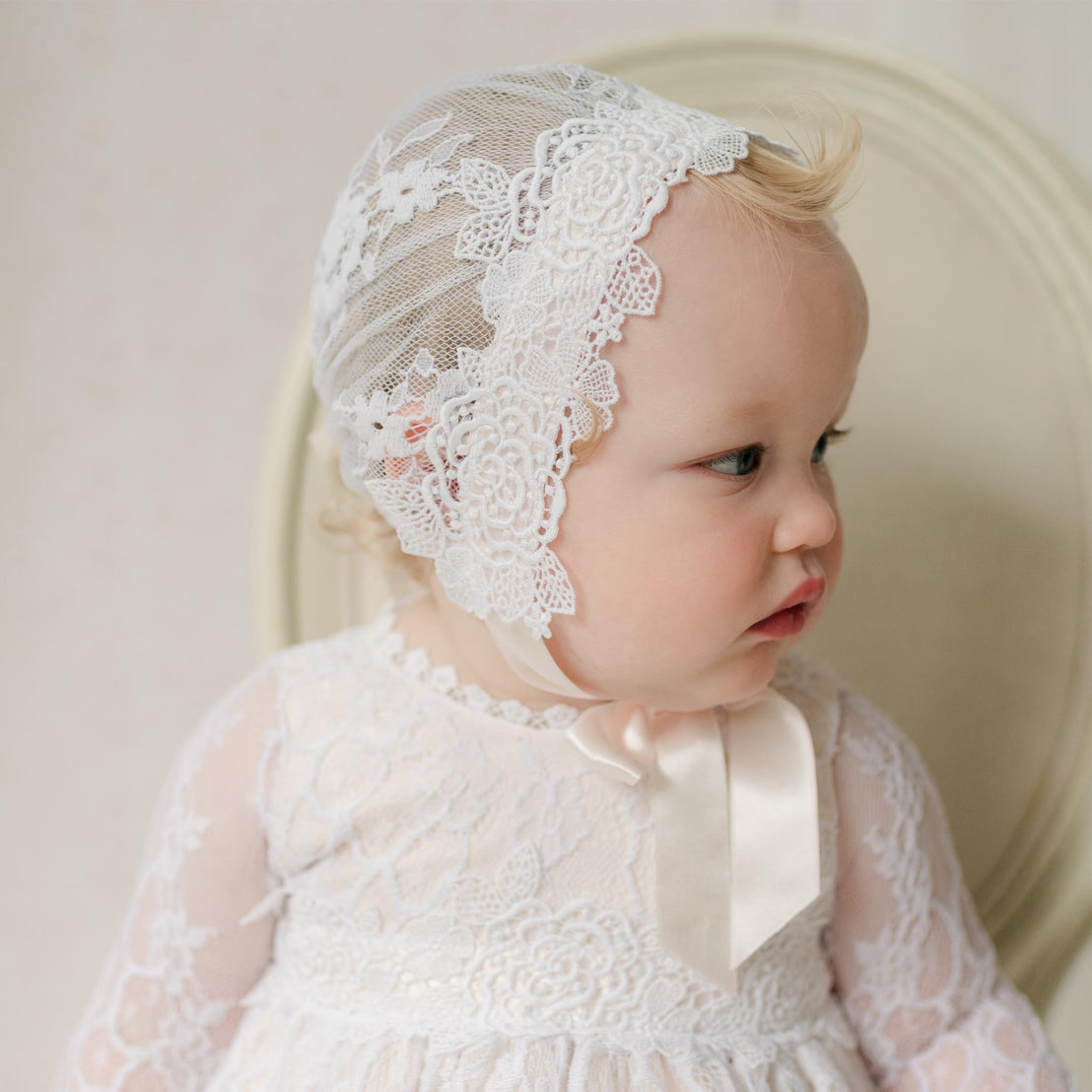 Baby wearing a white lace bonnet and dress against a neutral background