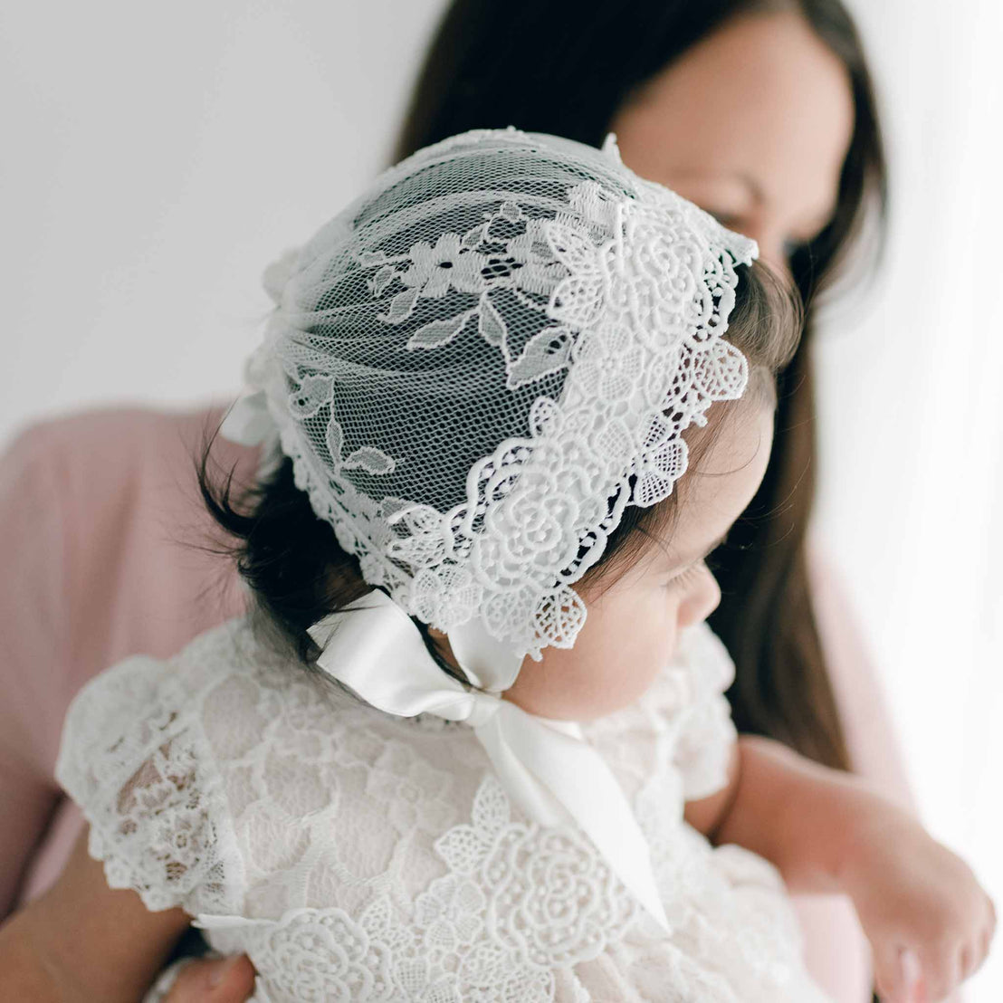 A close-up of a baby wearing a Juliette Lace Bonnet, cradled gently by her mother. This side profile highlights the floral lace and silk ribbon ties of the bonnet.