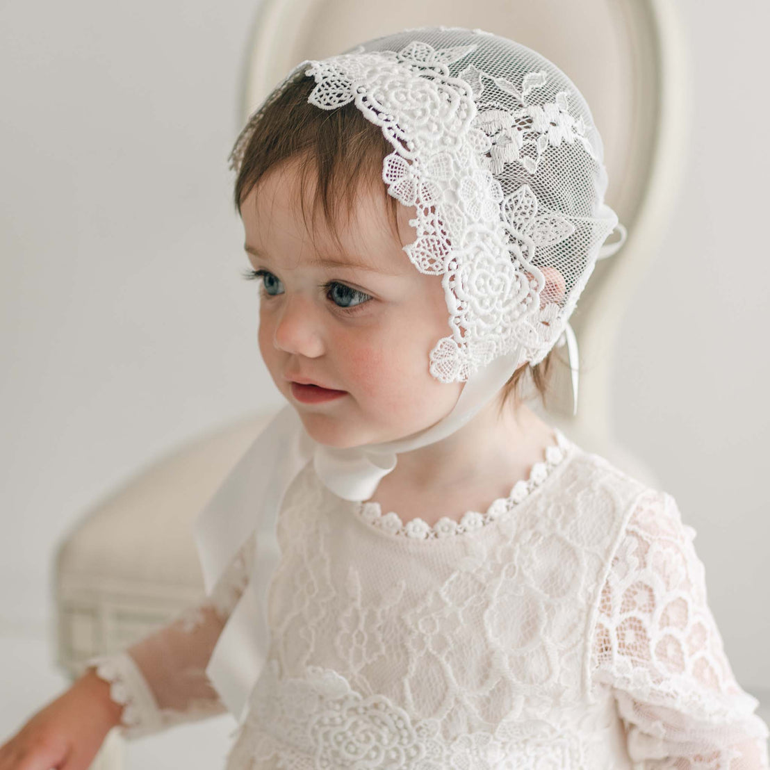 Baby girl wearing the Juliette Lace Bonnet adorned with intricate lace patterns and ivory silk ribbons, paired with a matching dress. She gazes to the side with an expression of curiosity.