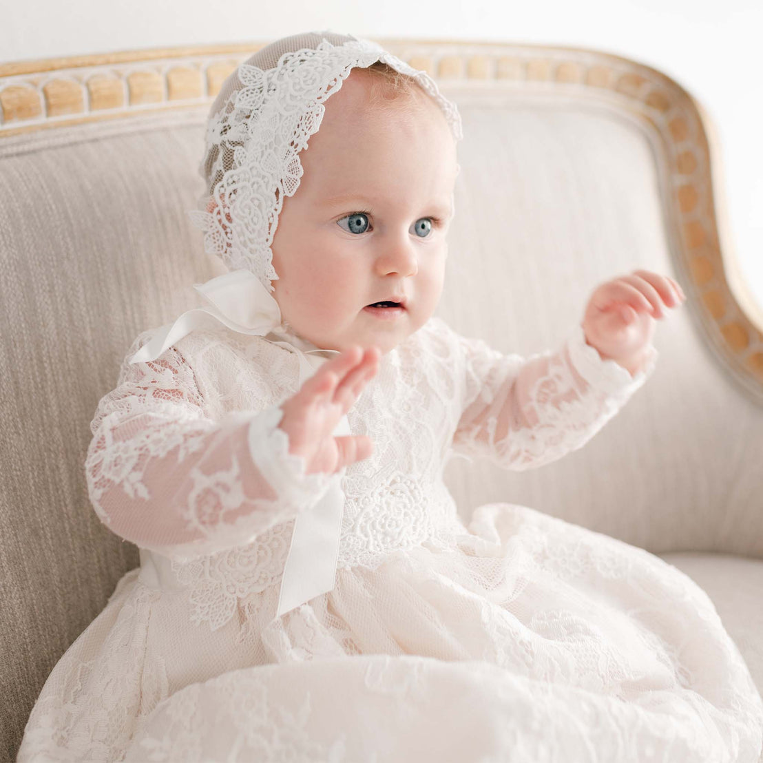 A baby girl in the Juliette Lace Bonnet and matching dress sits on a beige sofa with a curious expression, her arms raised in the air.