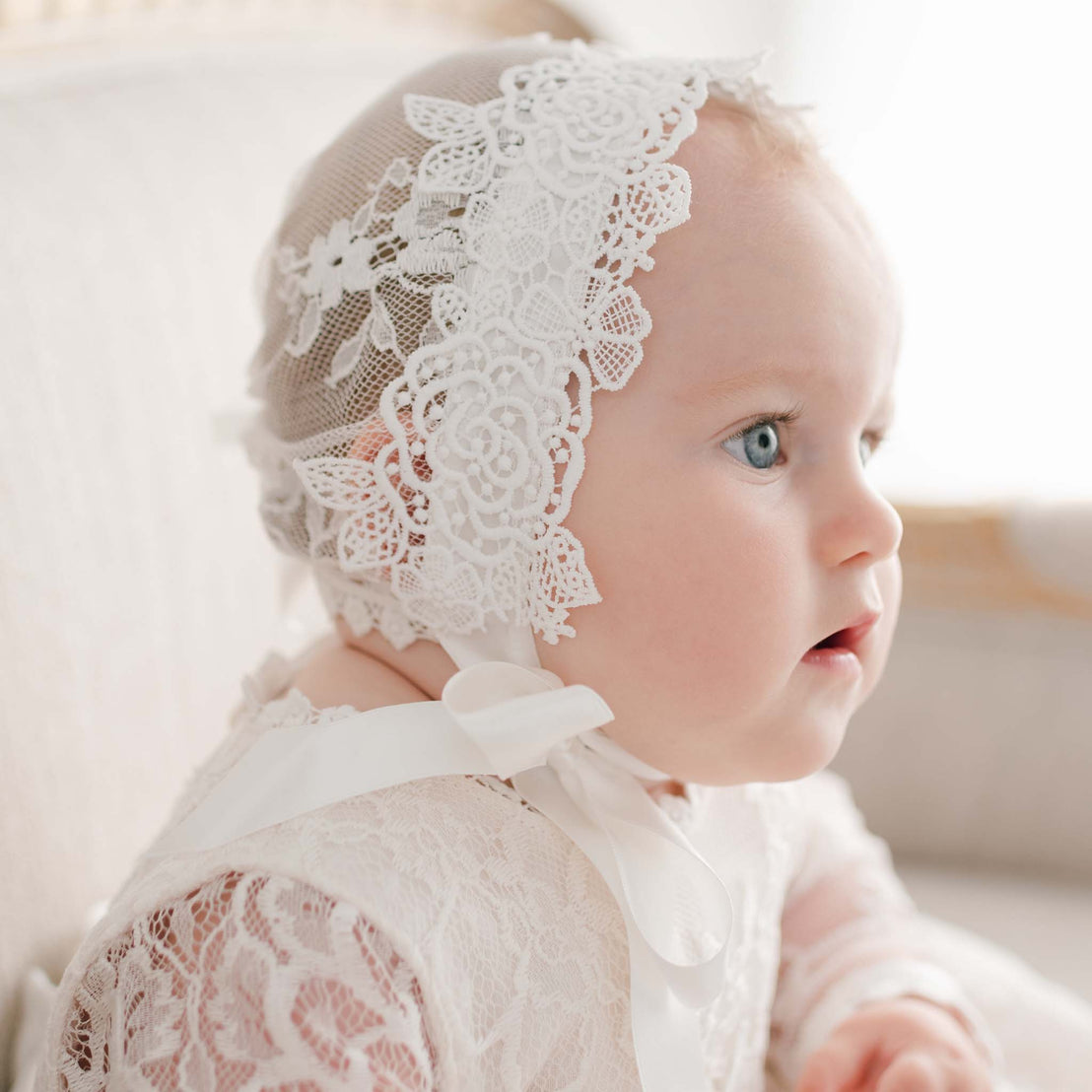 Side profile of a baby girl wearing the Juliette Lace Bonnet, featuring intricate floral lace and soft silk ribbon ties. The delicate lace detailing beautifully frames her face as she gazes off in the distance.