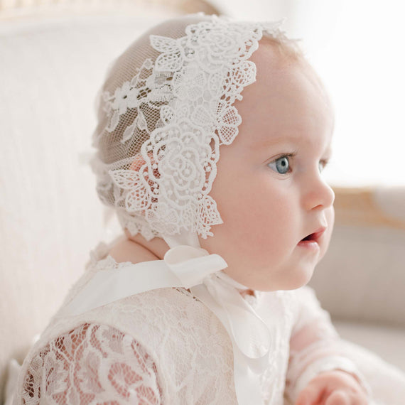 Side profile of a baby girl wearing the Juliette Lace Bonnet, featuring intricate floral lace and soft silk ribbon ties. The delicate lace detailing beautifully frames her face as she gazes off in the distance.