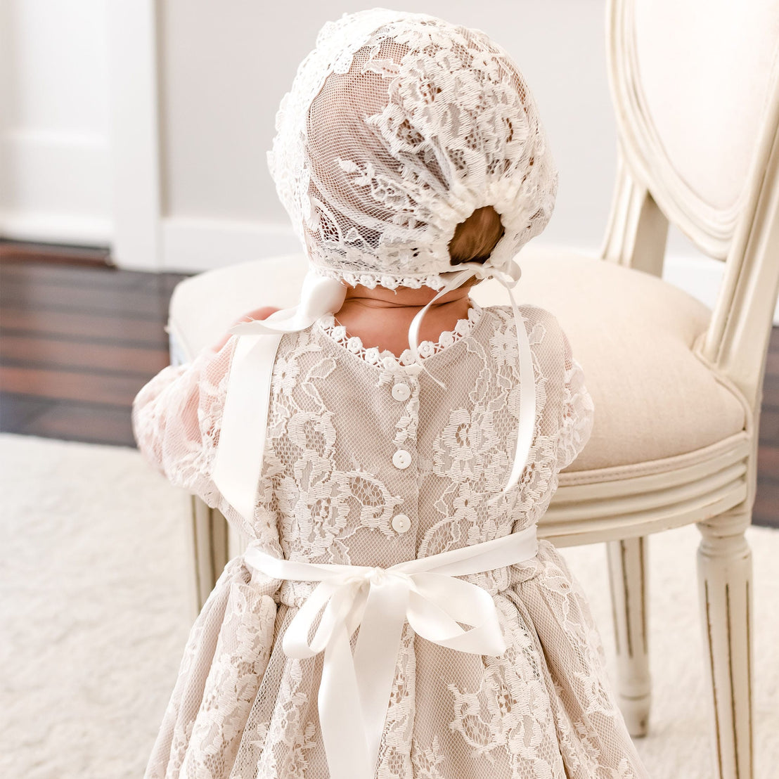 A baby viewed from behind wearing a cream lace dress and matching lace bonnet with white ribbon ties, standing indoors next to a light-colored upholstered chair.