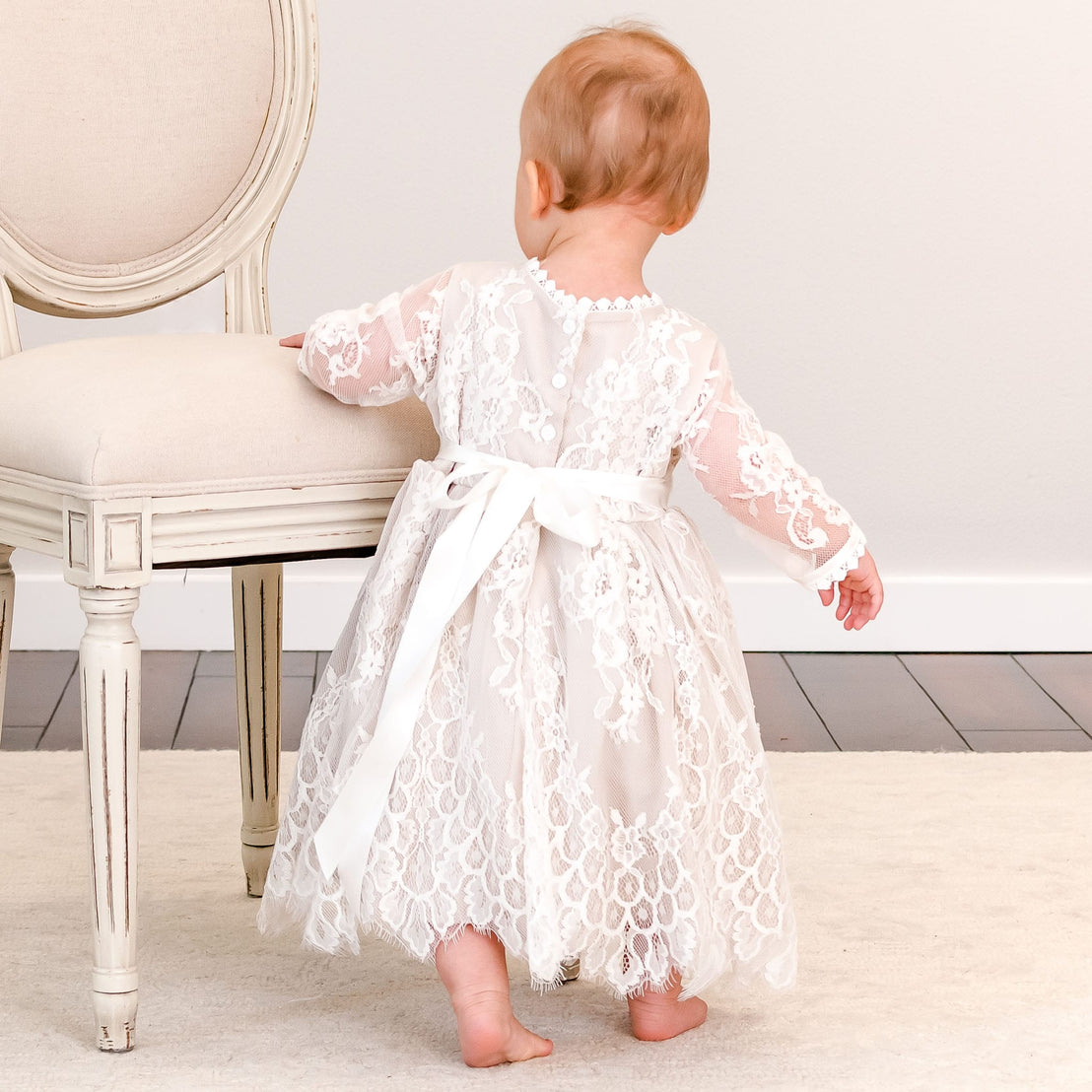 A baby girl seen from behind wearing a white lace dress with sheer sleeves and a matching white ribbon belt, standing on a light rug next to a vintage-style chair.