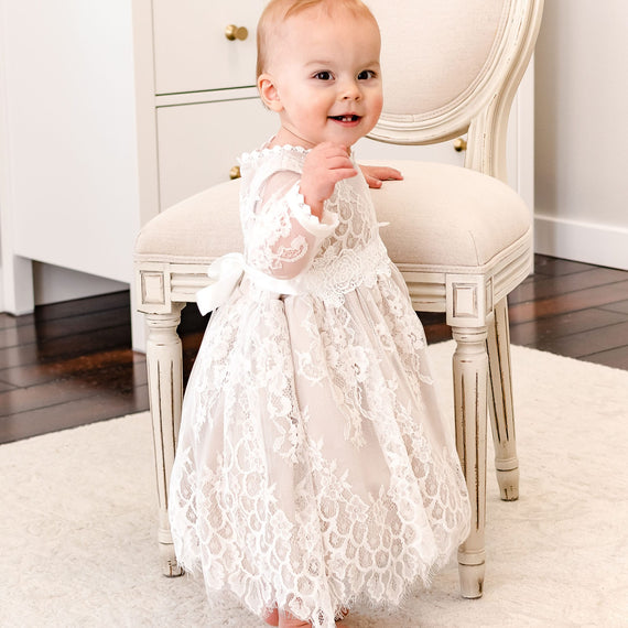 A baby girl wearing a long-sleeve white lace christening dress stands barefoot on a white rug in front of a cream-colored upholstered chair.