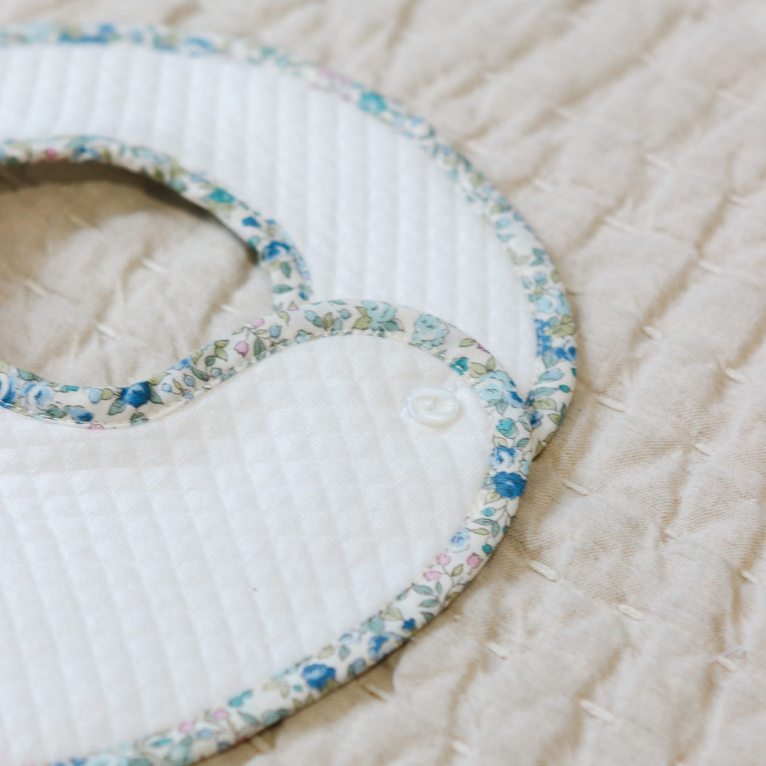Close up flat lay photo of the Lillian Bib on a beige surface. Close details show the blue floral cotton trim and button closure.