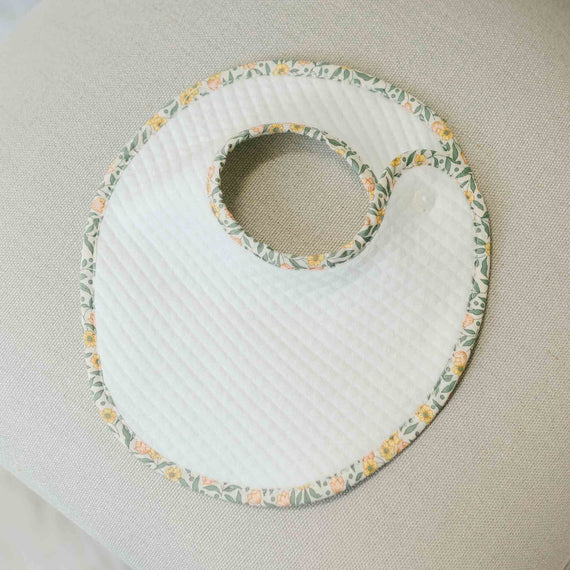 Flat lay photo of the Lillian Bib with floral cotton trim in yellow on a beige surface. Perfect for unique baby gifts for baby girl. 