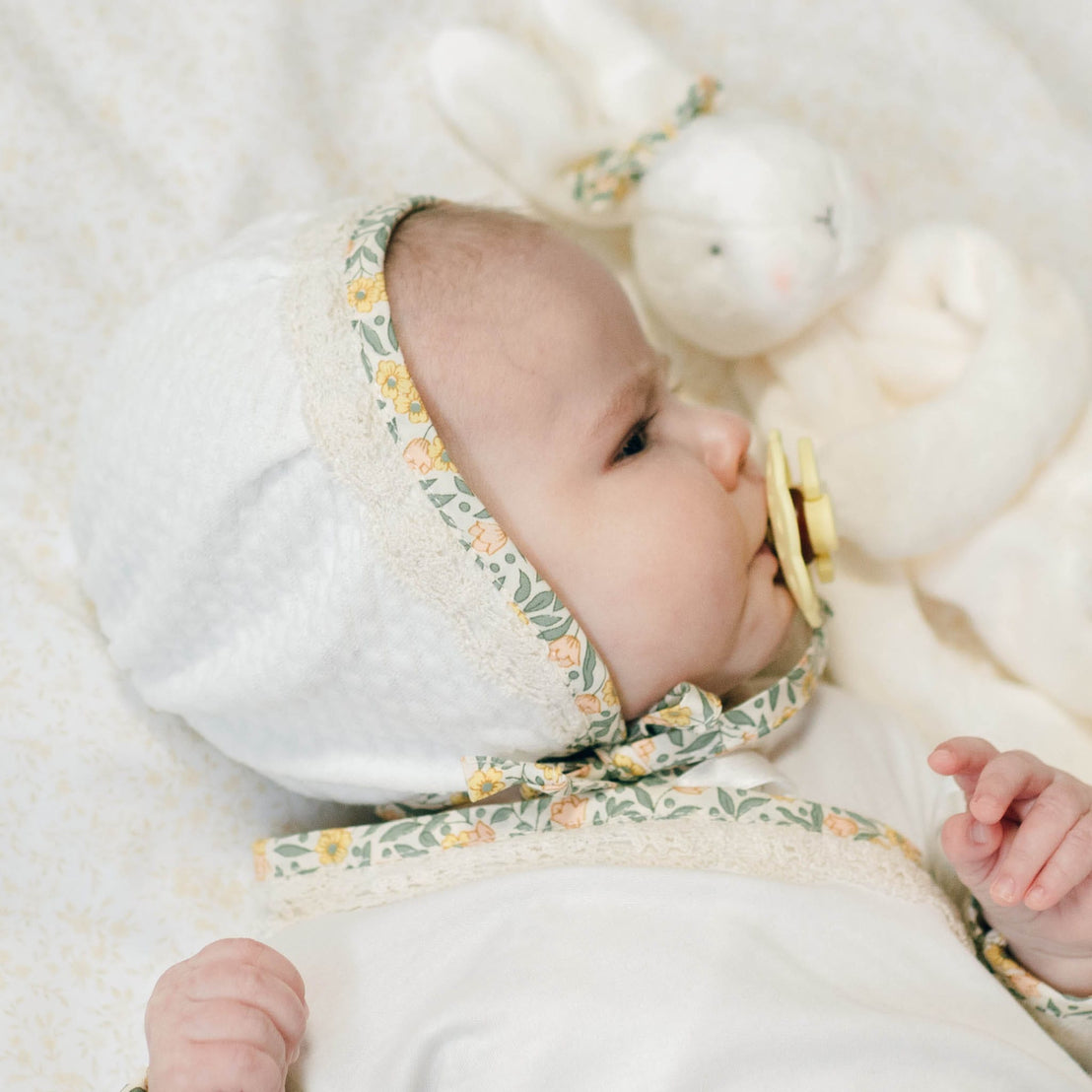 Baby in the Lillian Quilted Bonnet with yellow floral trim and matching Layette with a soft, blurred background. Perfect as a coming home outfit.