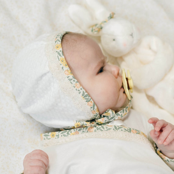 Baby in the Lillian Quilted Bonnet with yellow floral trim and matching Layette with a soft, blurred background. Perfect as a coming home outfit.