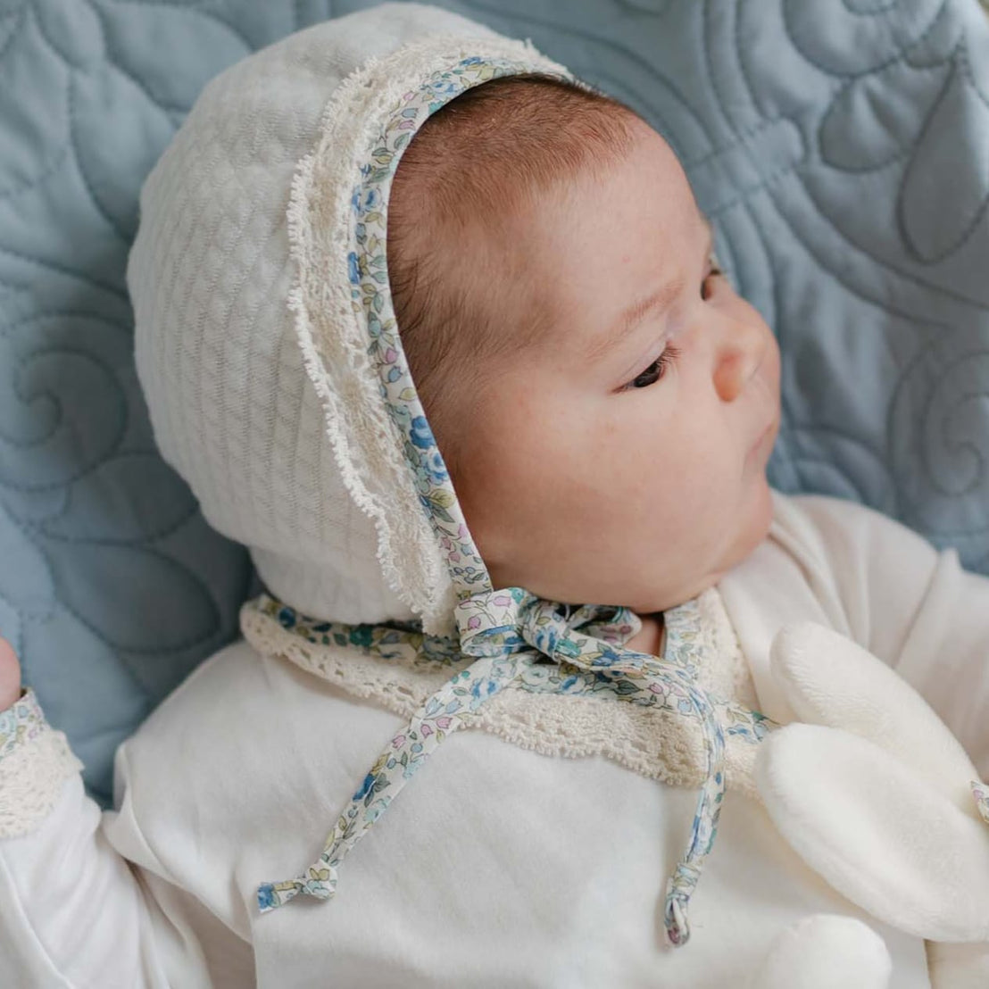 Baby lying in a wicker bassinet on a blue quilt, wearing the Petite Fleur Quilted Bonnet with floral trim and ecru lace, holding a soft bunny toy.