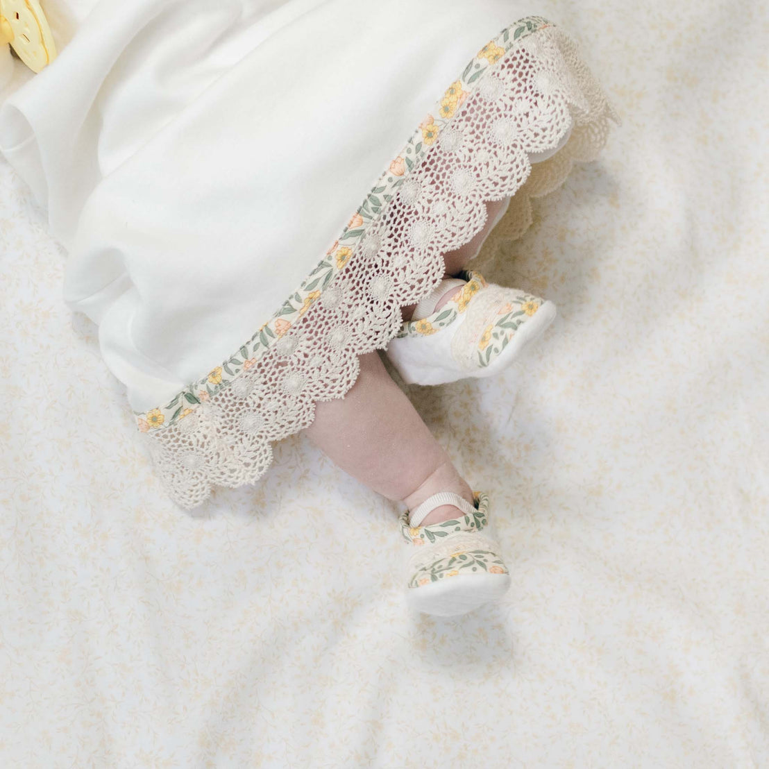 Baby's feet in the Lillian Booties peeking out from underneath the matching layette skirt. Set against a soft floral background.