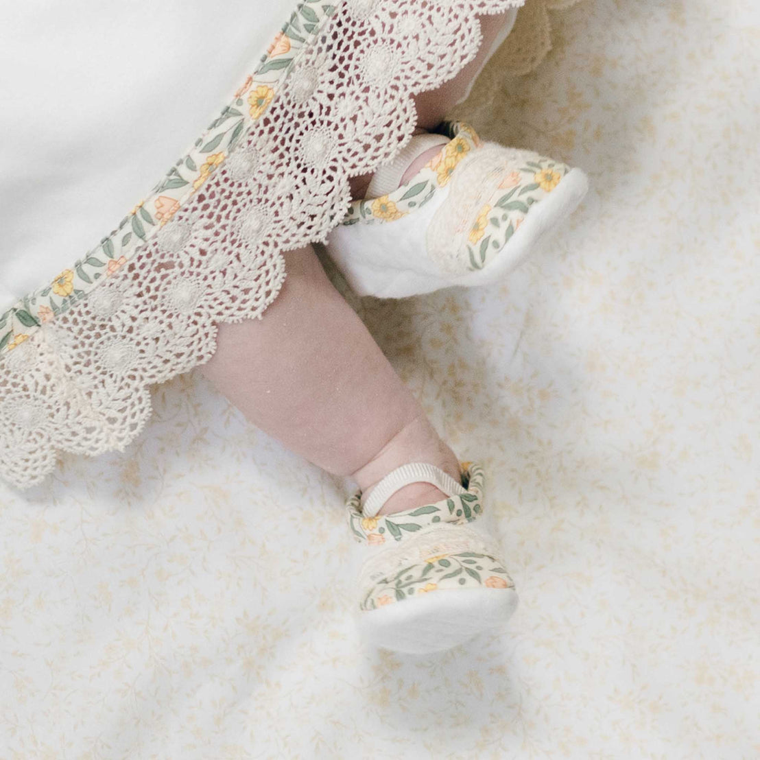 Close up photo of a baby's feet in the Lillian Booties peeking out from underneath the matching layette skirt. Set against a soft floral background.