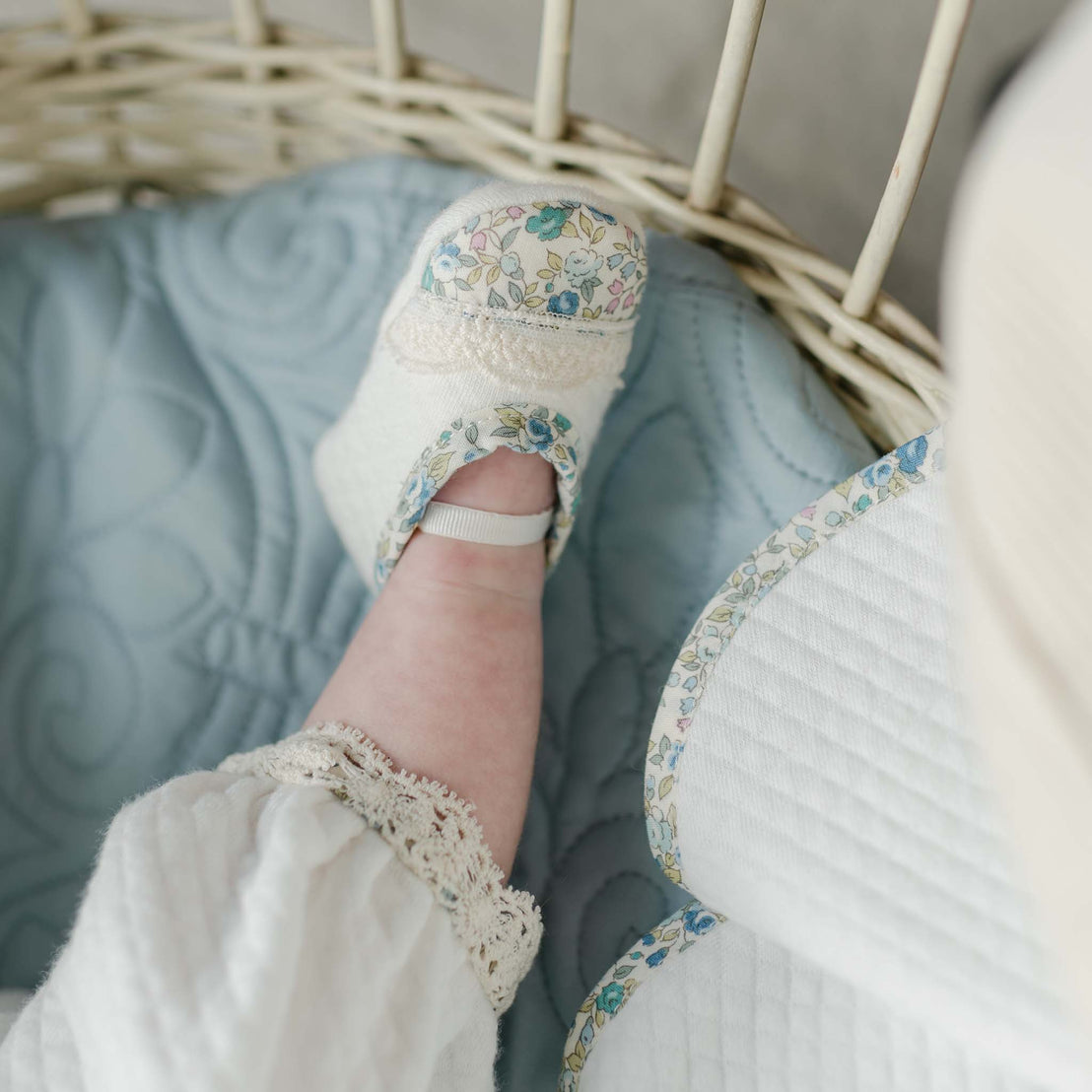 A close up photo of one of the Lillian Booties as worn by a baby in a wicker bassinet. Details show the blue floral trim and ivory lace. 