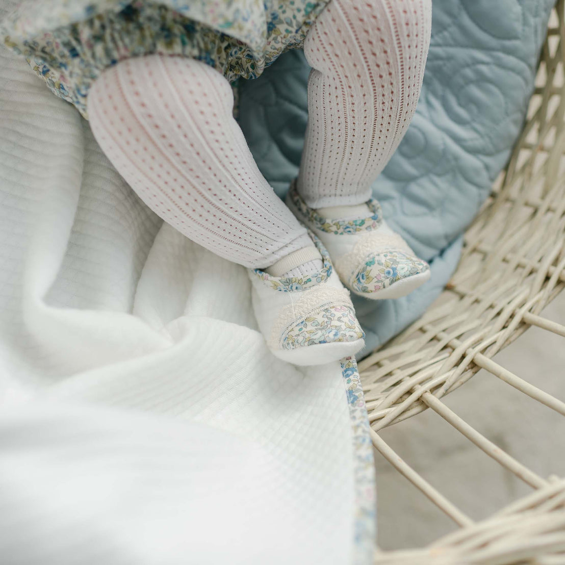 A pair of Lillian Booties with blue floral trim, worn by a baby in a wicker bassinet. Part of Baby Beau & Belle's high end baby clothes.