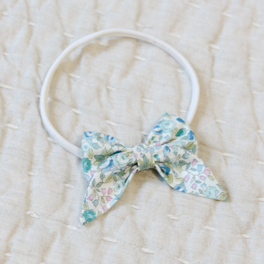 Close up flat lay photo of the Lillian Bow Headband set against a beige linen background. Details show the bow in a blue floral pattern. Perfect for unique baby gifts.