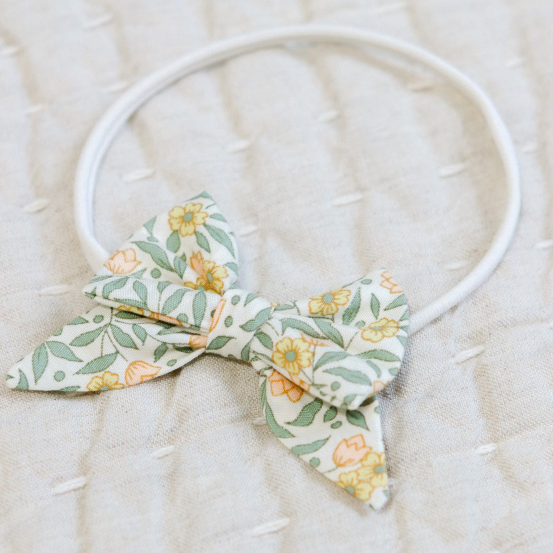 Close up flat lay photo of the Lillian Bow Headband set against a beige linen background. Details show the bow in a yellow floral pattern. Perfect for unique baby gifts.