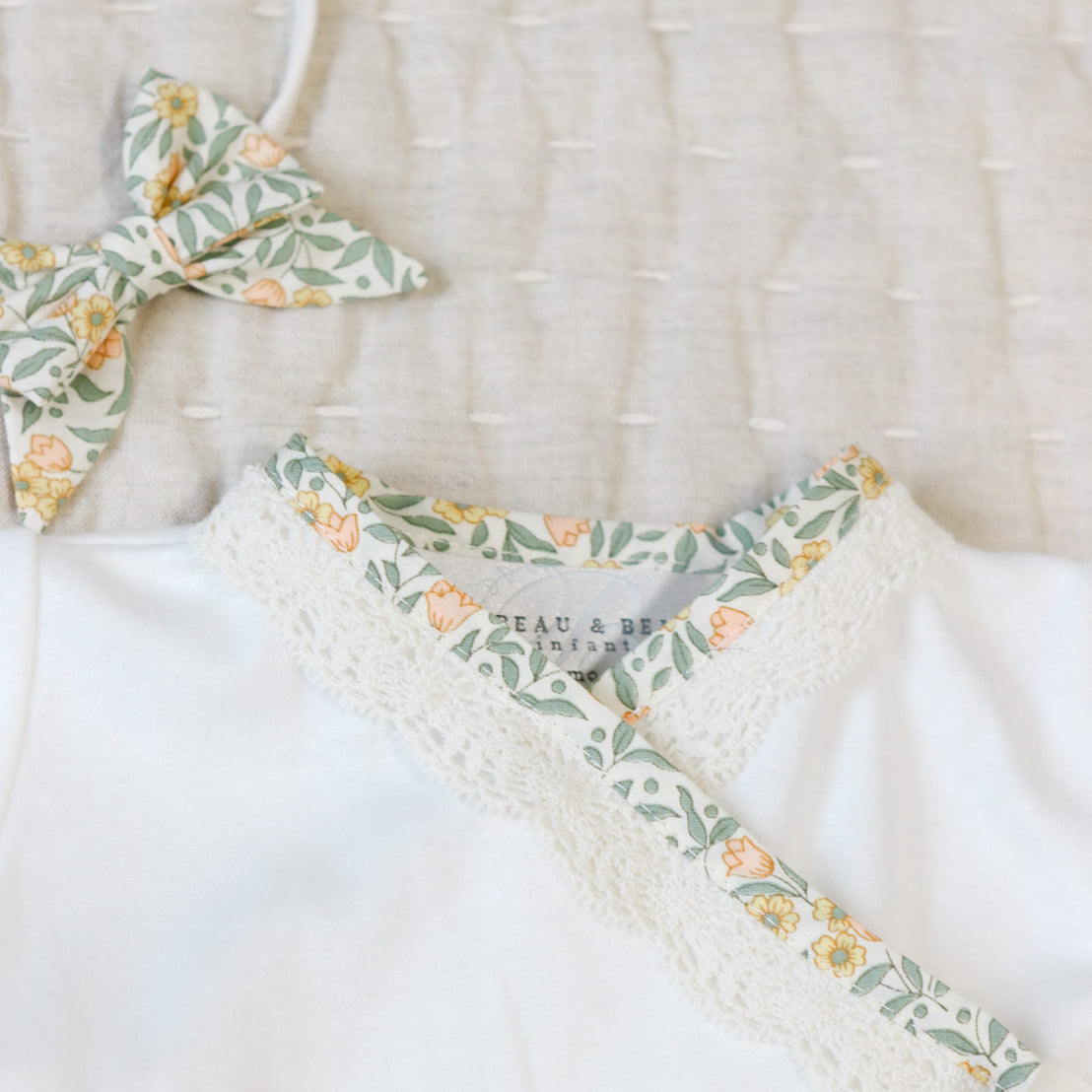 Close up flat lay photo of the Lillian Layette with yellow floral cotton and scalloped champagne lace trim. Set against a beige linen background.