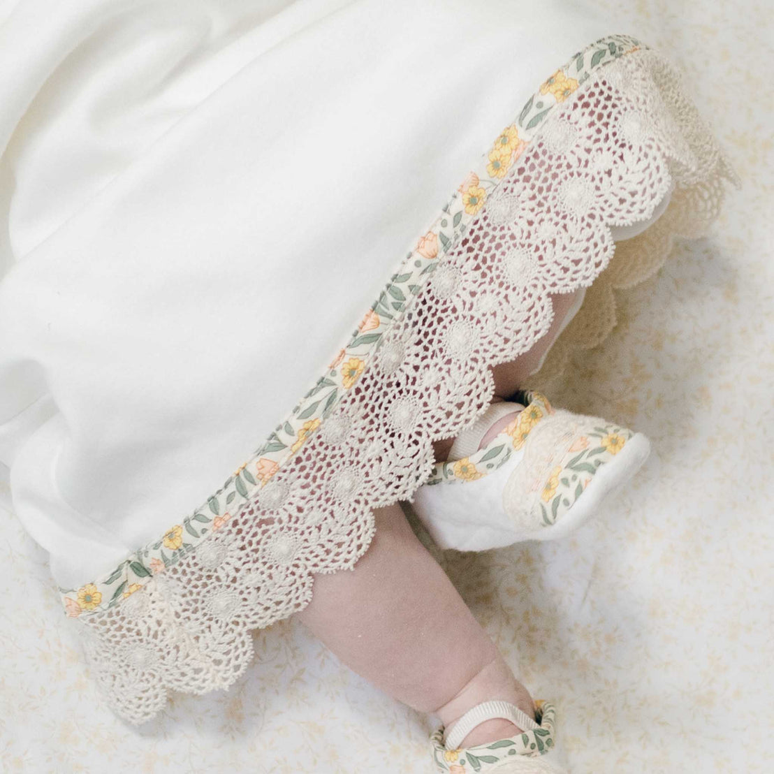 Close up photo of a baby's feet in the Lillian Booties and peeking out from the matching layette skirt. Details show the scalloped champagne lace that lines the skirt. Part of Baby Beau & Belle's Coming Home Outfit Collection.